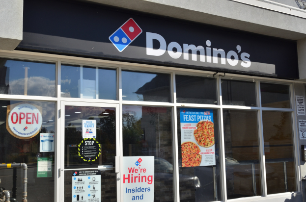 Außenansicht eines Domino's Pizza-Restaurants mit Glaswänden und -türen, das Stellenanzeigen und ein Namensschild an der Wand zeigt.