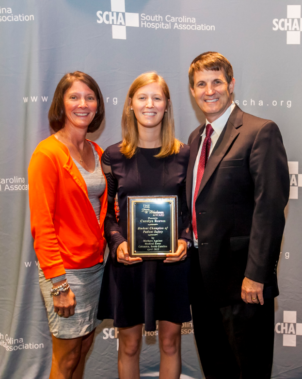 Drei Menschen, zwei Frauen und ein Mann, stehen zusammen. Der Mann trägt einen schwarzen Anzug, eine Frau trägt ein orange-schwarzes Kleid und die andere Frau trägt ein schwarzes Kleid und hält eine Trophäe in der Hand.