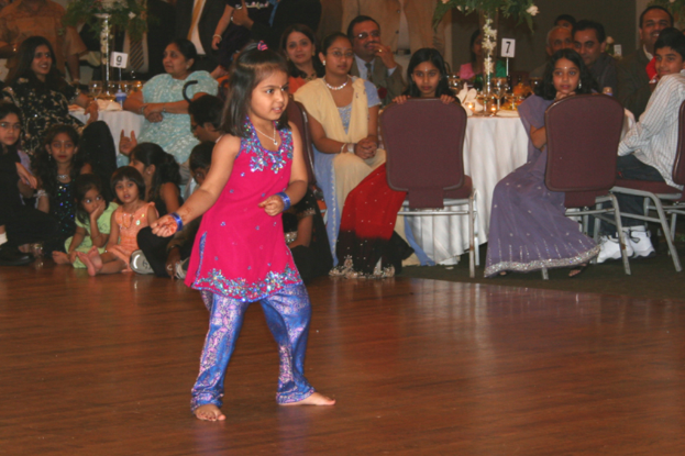 Ein Mädchen tanzt im Vordergrund mit einem Holzfußboden darunter, während im Hintergrund viele Menschen an Tischen sitzen, die mit weißen Tischdecken und kleinen Pflanzen bedeckt sind.