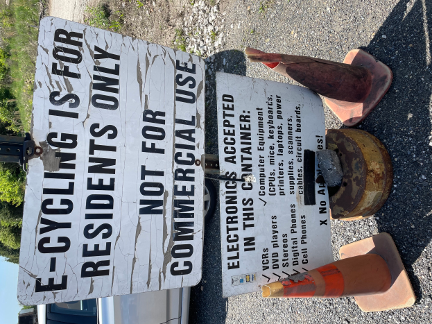 Ein Schild mit der Aufschrift "E-Bike-Fahren nur für Anwohner" wird an der Seite einer Straße aufgestellt, umgeben von Verkehrskegeln und anderen Gegenständen, mit einem Fahrzeug, Bäumen und einem klaren blauen Himmel im Hintergrund.