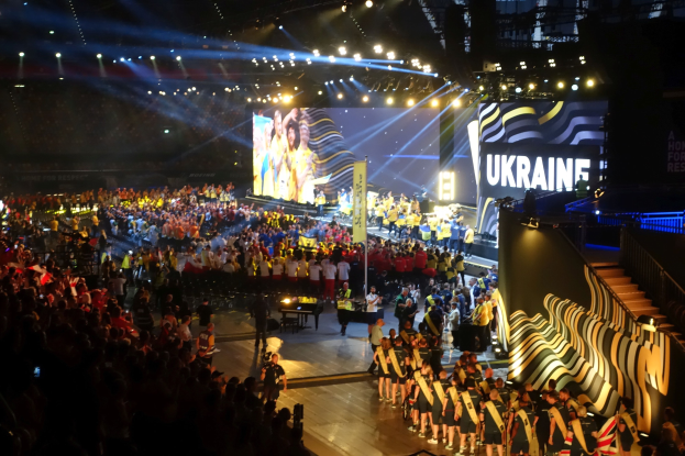 Eine große Menschenmenge auf einer hell erleuchteten Bühne mit Musikern, die Instrumente spielen, und einem Bildschirm im Hintergrund, der auf ein bevorstehendes Ereignis hinweist.