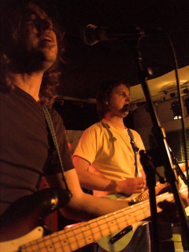 Eine Person in einem wei├čen Hemd singt in ein Mikrofon und spielt eine gr├╝ne Gitarre, steht neben einer anderen Person in einem schwarzen Hemd und Brille, die ebenfalls singt und eine schwarze Gitarre spielt, mit Scheinwerfern und anderen Instrumenten im Hintergrund.