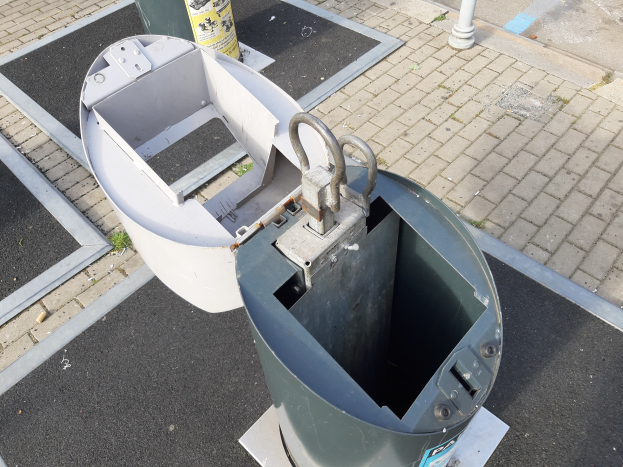 Ein Metallmüllcontainer an der Seite einer Straße, mit einem Metallobjekt im Vordergrund und einem Pfahl im Hintergrund.