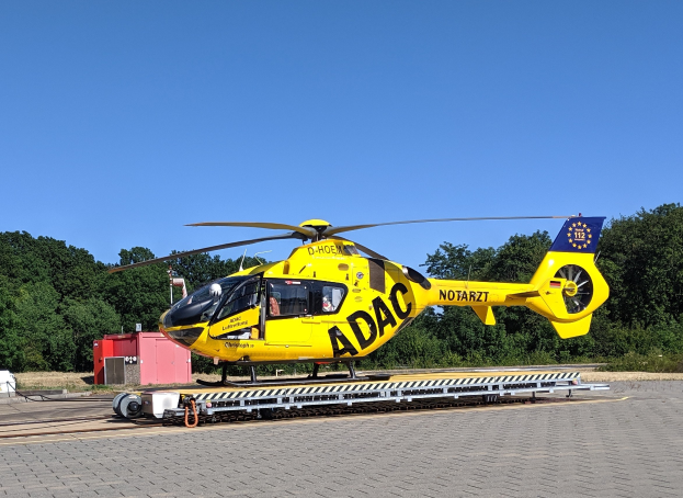 Gelber Hubschrauber auf einem Flughafenvorfeld mit umliegenden Bäumen und einem klaren blauen Himmel, wahrscheinlich für Luftrettungsdienste in Deutschland verwendet.