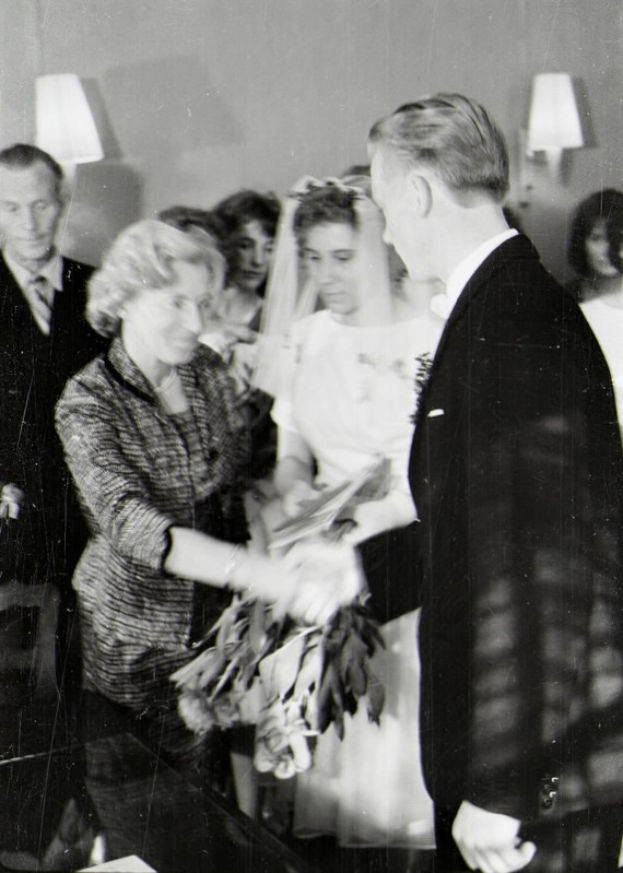 Ein Schwarz-Weiß-Foto eines Brautpaares, das sich die Ringe austauscht, mit der Braut in einem weißen Kleid und Schleier, die einen Strauß hält, und dem Bräutigam in einem Anzug und Krawatte, mit Wandlampen im Hintergrund.