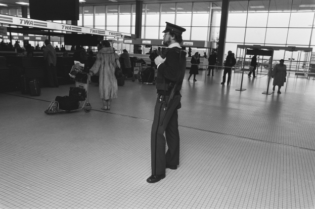 Polizeibeamter in Uniform mit Mütze, der eine Waffe hält und in einem Flughafen-Terminal umgeben von Menschen und Gepäck steht, mit Gepäckwagen, Deckenleuchten, Pfosten, Glaswänden und Informationsanzeigen im Hintergrund.