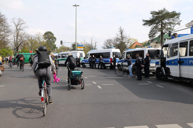 Gruppe von Menschen auf Fahrrädern auf einer Straße mit Polizeiwagen, anderen Fahrzeugen, Bäumen, Laternen, Verkehrsampeln, Verkehrsschildern, Gebäuden und einem bewölkten Himmel.