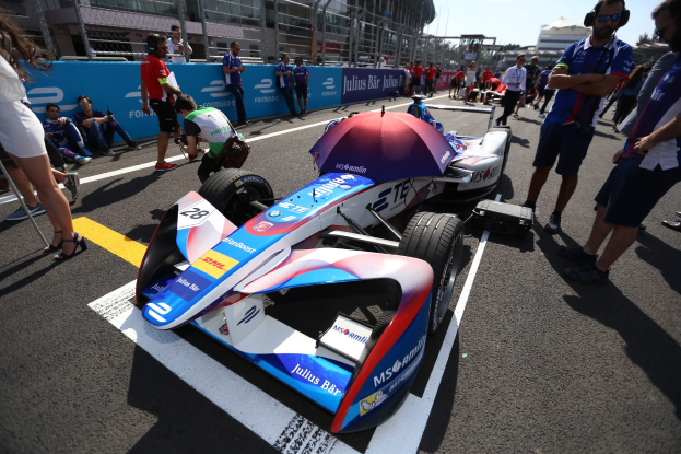 Formula E-Auto mit weißer, blauer, roter und schwarzer Lackierung auf einer Rennstrecke geparkt, umgeben von Menschen, mit einem Schirm im Vordergrund und Rennstreckenabsperrungen, Gebäuden, Bäumen und einem klaren blauen Himmel im Hintergrund.
