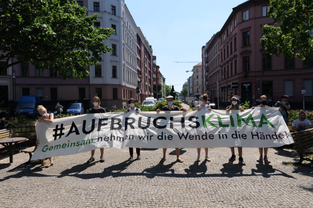 Eine Gruppe maskierter Personen mit einem Banner "Aufbruchsklima" protestiert gegen den Klimawandel in Berlin, Deutschland, mit Gebäuden, Bänken, Tischen, Pflanzen, Bäumen, Fahrzeugen, Laternenmasten, Schildern und anderen Menschen im Hintergrund.