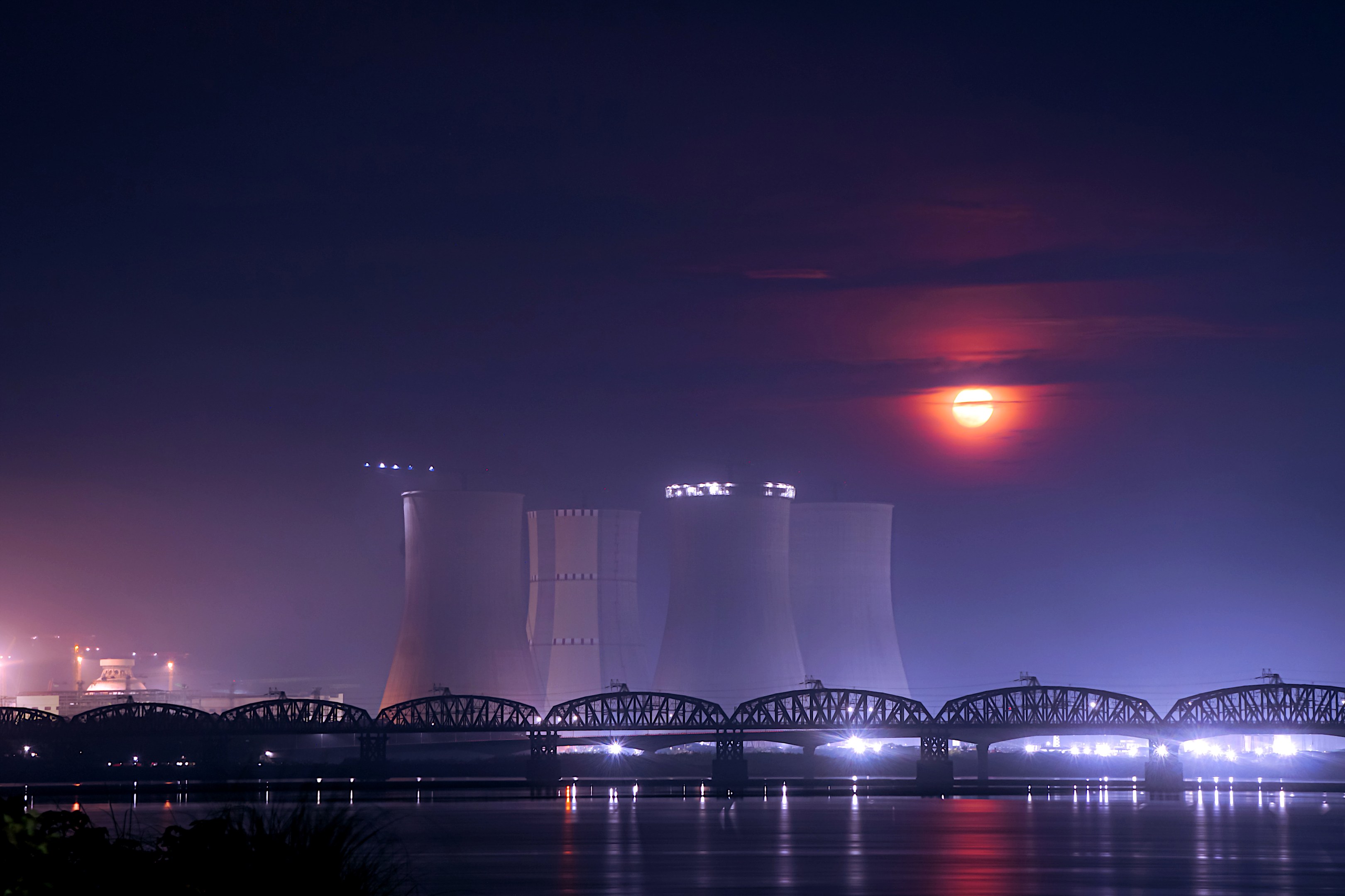 Ein Kernkraftwerk bei Nacht, mit einer Brücke über Wasser, beleuchtet von Gebäuden und Mondlicht, umgeben von Bäumen.