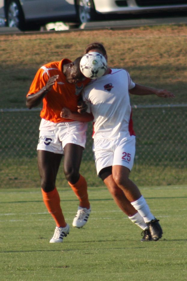 Zwei Spieler auf einem umzäunten Platz, einer im orangen T-Shirt, der den Ball mit dem Kopf schlägt, mit Autos und Gras im Hintergrund.