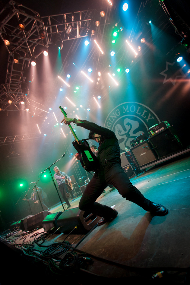 Ein musikalisches Konzert mit drei Personen auf der Bühne: zwei spielen rechts Gitarre mit Mikrofonen davor, ein Hintergrund mit "MOLLEY" geschrieben und zwei Schlangenlogos, sowie Bühnenlichter oben.