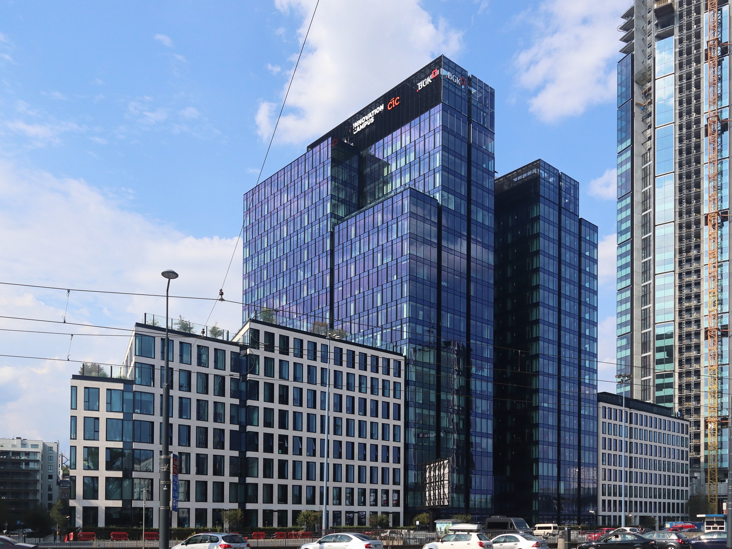 Außenansicht der Deutschen Bank Hauptverwaltung in Frankfurt, Deutschland, mit einem großen Gebäude mit vielen Fenstern, umgeben von städtischer Infrastruktur wie Straßenlaternen, Stromkabeln, Fahrzeugen, Grünflächen und einem bewölkten Himmel.