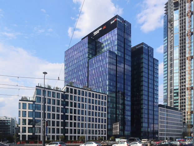 Außenansicht der Deutschen Bank Hauptverwaltung in Frankfurt, Deutschland, mit einem großen Gebäude mit vielen Fenstern, umgeben von städtischer Infrastruktur wie Straßenlaternen, Stromkabeln, Fahrzeugen, Grünflächen und einem bewölkten Himmel.