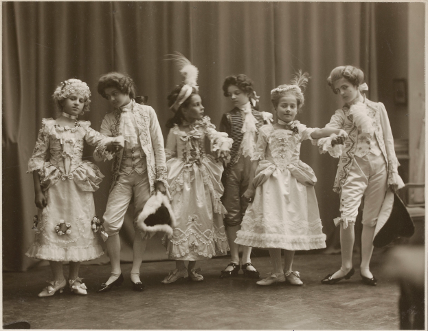 Eine Gruppe von Kindern in Kleidern und Schuhen steht zusammen auf einer Bühne mit einem Vorhang im Hintergrund.