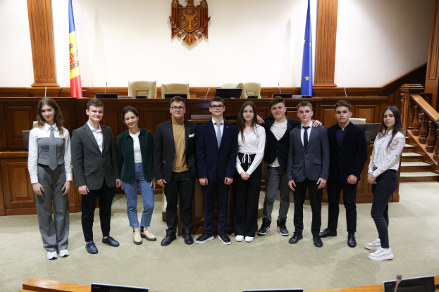 Gruppe von strahlenden Menschen, die vor einem Gerichtssaal mit Monitoren und Mikrofonen auf Tischen, einer hölzernen Plattform mit Stühlen und Fahnen im Hintergrund und einer Treppe auf der rechten Seite posieren.