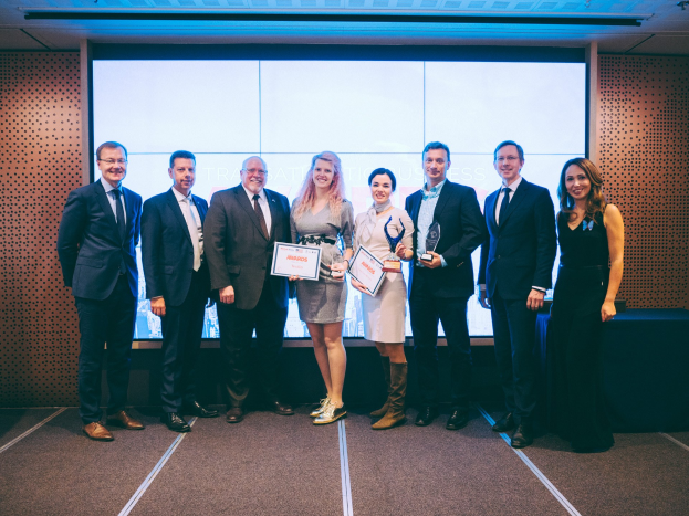 Eine Gruppe strahlender Menschen mit Urkunden, die vor einem Bildschirm mit einem Tisch und einem wandmontierten Objekt im Hintergrund stehen - Gewinner der Internationalen Business Awards 2018.