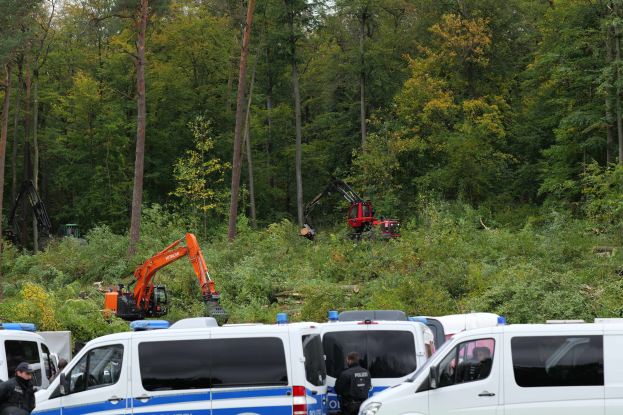 Eine Gruppe von Polizeiwagen in einem waldigen Gebiet geparkt, mit Officers und anderen in der Nähe, ein Bagger im Hintergrund und Täler in der Umgebung unter einem offenen Himmel.