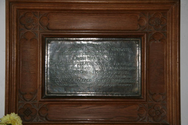 Eine Gedenktafel mit Text ist an einer braunen Wand oder einer Holztür angebracht, mit einem gelben Objekt, das einer Vase in der linken unteren Ecke ähnelt.