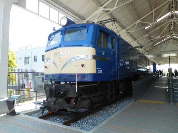 Ein blau-weißer Zug fährt auf Schienen neben einem Bahnsteig mit ein paar Menschen, einer Treppe auf der rechten Seite und einem Geländer auf der linken Seite, mit Bäumen, einem Gebäude und dem Himmel im Hintergrund.