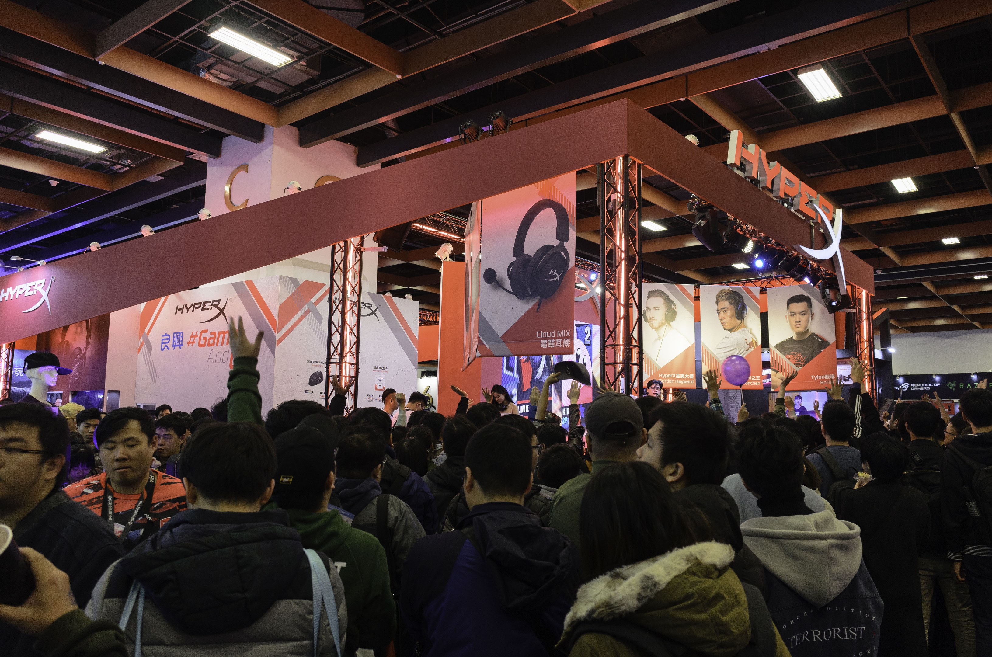 Eine große Menschenmenge steht vor einem Stand auf einer Gaming-Messe in China, einige tragen Taschen und einer hält einen Becher; Banner und Deckenleuchten sind im Hintergrund zu sehen.