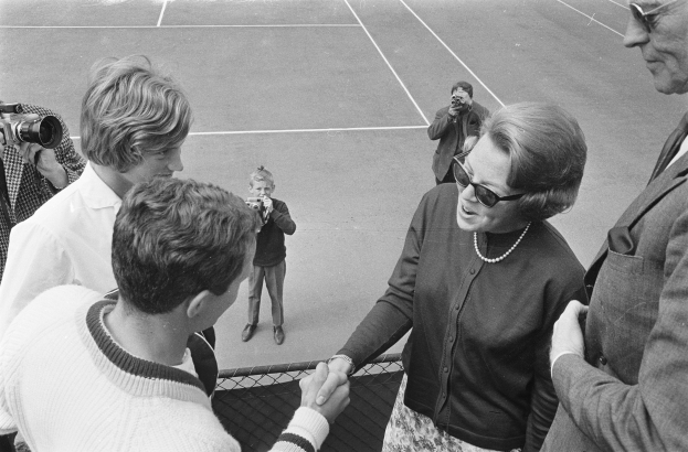 Eine Gruppe von Menschen auf einem Tennisplatz, mit einer Frau in einer Brille, die einem Mann im Anzug die Hand sch├Ąktelt, und Fotografen im Hintergrund, alles in Schwarz-Wei├č.