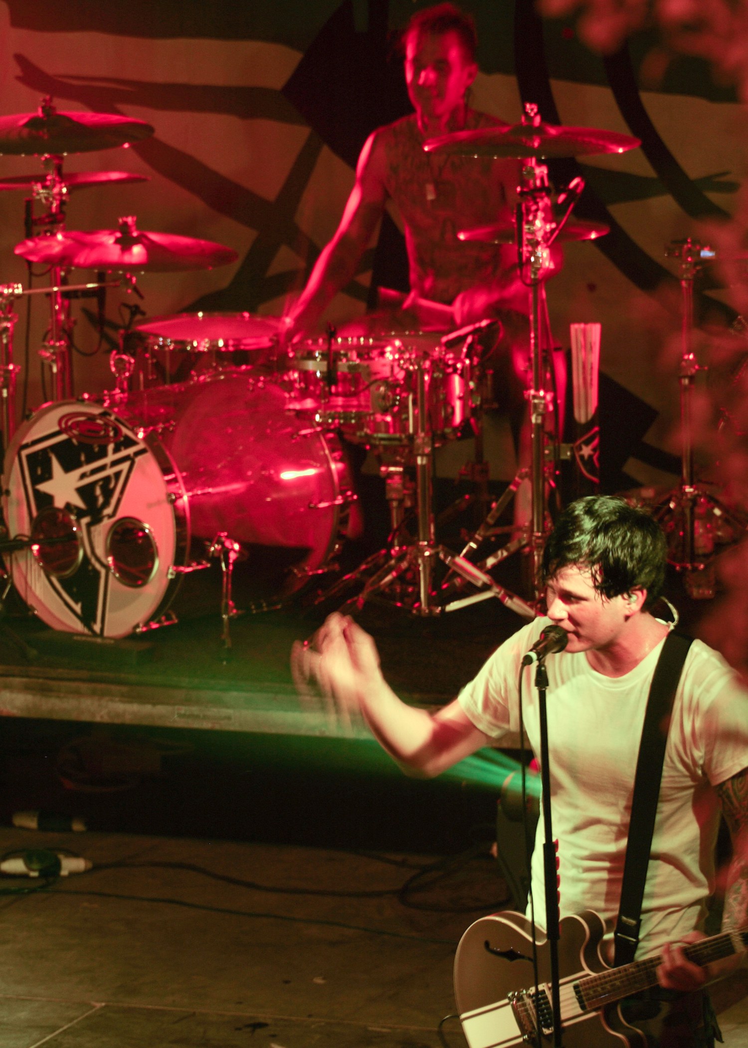 Ein Mann, der auf der Bühne eine E-Gitarre spielt, mit einem Mikrofon davor und einer Person, die im Hintergrund Schlagzeug spielt.