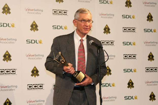 Ein Mann mit Brille steht vor einem Mikrofon und hält eine Trophäe in der Hand, mit einem Schild im Hintergrund.