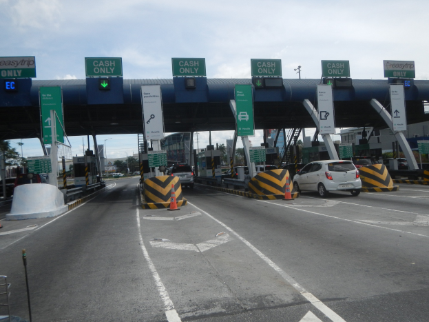 Mautstation mit darunter fahrenden Autos, umgeben von Verkehrskegeln, Absperrpoller, Schildern, einem Bogen, Laternenmasten, Bäumen, Gebäuden und einem bewölkten Himmel.
