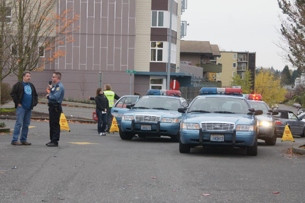 Autos auf einer Straße mit vier Menschen in der Nähe, Gebäude mit Fenstern im Hintergrund, Bäume und Warnwesten.