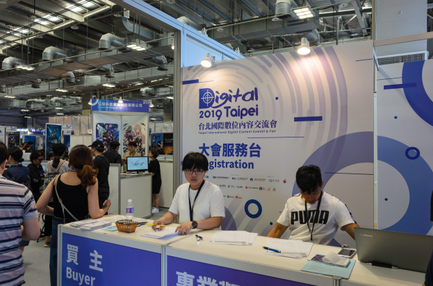 Eine Gruppe von Menschen steht um einen Stand auf einer Messe, mit zwei Personen, die an einem Tisch mit verschiedenen Gegenständen sitzen, und Bannern mit Text im Hintergrund, die auf die Digital 2019 Taipei International Digital Content Summit & Fair hinweisen.