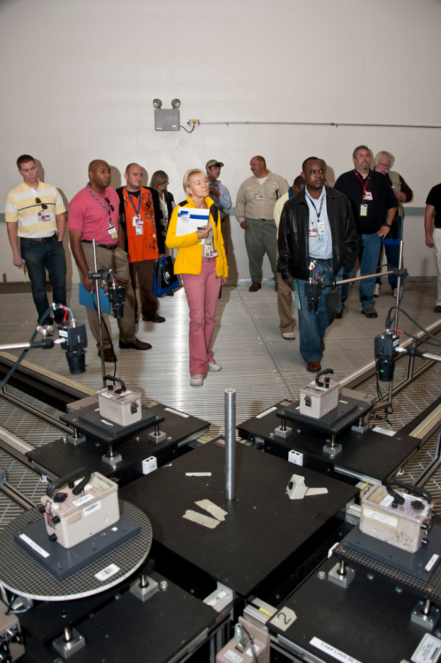 Eine Gruppe von Menschen in einem Raum, die ID-Karten tragen und Ordner und Kameras halten, vor verschiedenen Geräten mit einer Wand im Hintergrund.