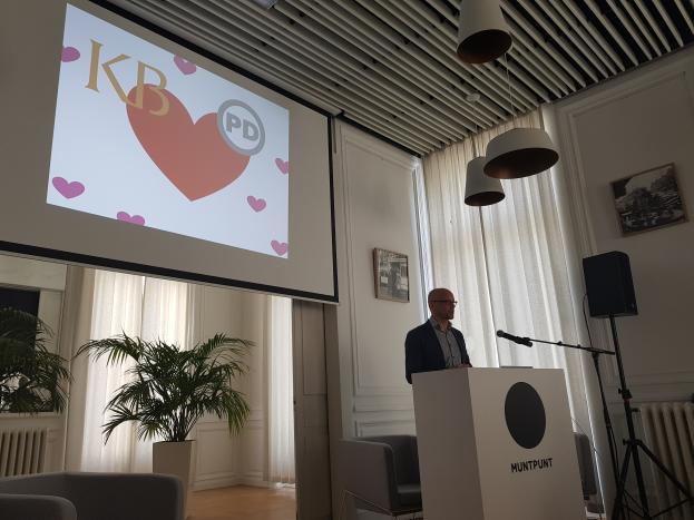 Ein Mann steht an einem Rednerpult mit einem Mikrofon und hält eine Präsentation vor einem Projektionsschirm, mit Stühlen, einer Pflanze, Fotorahmen, Vorhängen und Deckenleuchten im Raum.