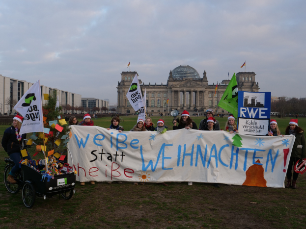 Eine Gruppe von Menschen mit Mützen und einem Banner vor dem Reichstaggebäude in Berlin, Deutschland, mit einer Person auf einem dekorierten Kinderwagen, Gras unter den Füßen, Bäumen und Gebäuden mit Fenstern im Hintergrund und Fahnen mit Stangen, unter einem bewölkten Himmel.