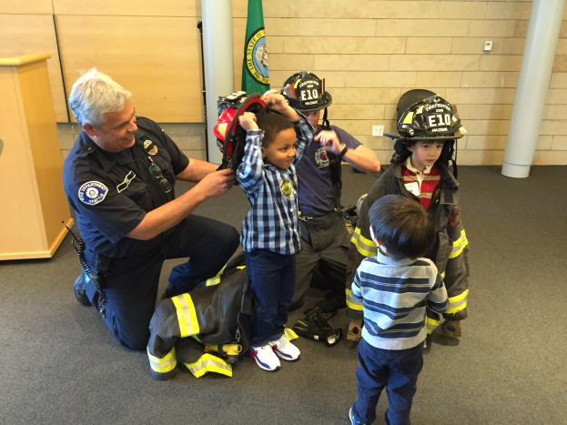 Eine Gruppe von Kindern in Feuerwehruniformen und ein Mann in einer Feuerwehruniform stehen neben einem Helm und einer Tasche in einer Feuerwache, mit Säulen, einer Flagge und einem hölzernen Objekt im Hintergrund.