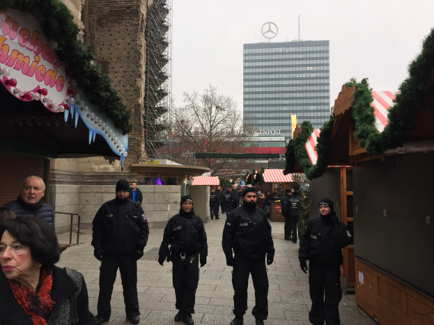 Eine Gruppe von Polizisten in schwarzen Uniformen steht vor einem festlichen Weihnachtsmarkt in Berlin, Deutschland, mit Gebäuden, Bäumen und einem klaren blauen Himmel im Hintergrund und einer Tafel mit Schrift auf der linken Seite des Bildes.