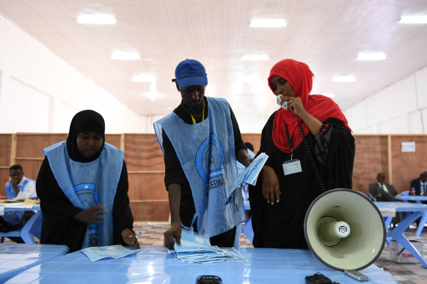 Eine Gruppe von Menschen steht um einen Tisch mit einem Megafon, Papieren und anderen Gegenständen herum, die scheinbar an einer Wahllokalität sind, mit anderen im Hintergrund auf Stühlen sitzend und stehend unter Deckenleuchten.