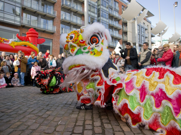 Ein lebhaftes chinesisches Neujahrsfest in Amsterdam mit einem Löwen tanzen und einer Menschenmenge, einige halten Kameras, vor einem Hintergrund aus Gebäuden, Laternenmasten und einem klaren blauen Himmel.