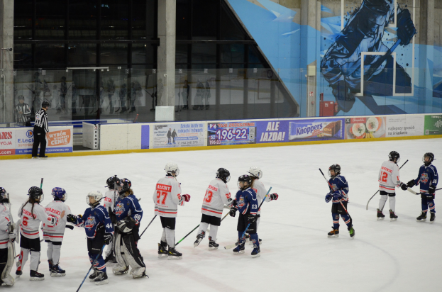 Gruppe junger Menschen, die Eis-hoc-key auf einer Indoor-Eisfläche spielen, mit Helmen und Stöcken, sowie einer Person auf der linken Seite und Bannern, Säulen und Glaswänden im Hintergrund.