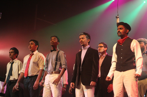 Eine Gruppe von Männern auf einer Bühne, die ein Drama mit grünen Scheinwerfern im Hintergrund aufführen.