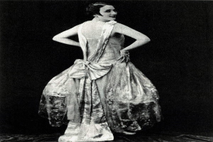 Eine Frau in einem Kleid im Stil der 1920er Jahre mit hohem Kragen, langen Ärmeln und einem weiten Rock steht selbstbewusst in einem Schwarz-Weiß-Foto, ihr Haar ist elegant hochgesteckt und das Make-up dezent.