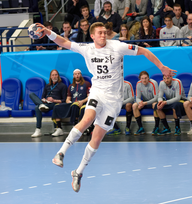 Ein Mann in der Luft, der einen Fußball hält, umgeben von Menschen auf Stühlen mit einem Geländer im Hintergrund, die Schuhe tragend.