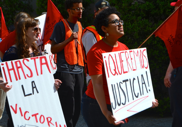 Mehrere Personen in orangefarbenen Kleidern stehen mit weißen Schildern und orangefarbenen Flaggen, eine Frau in einem schwarzen Kleid steht im Vordergrund und grüne Bäume im Hintergrund.