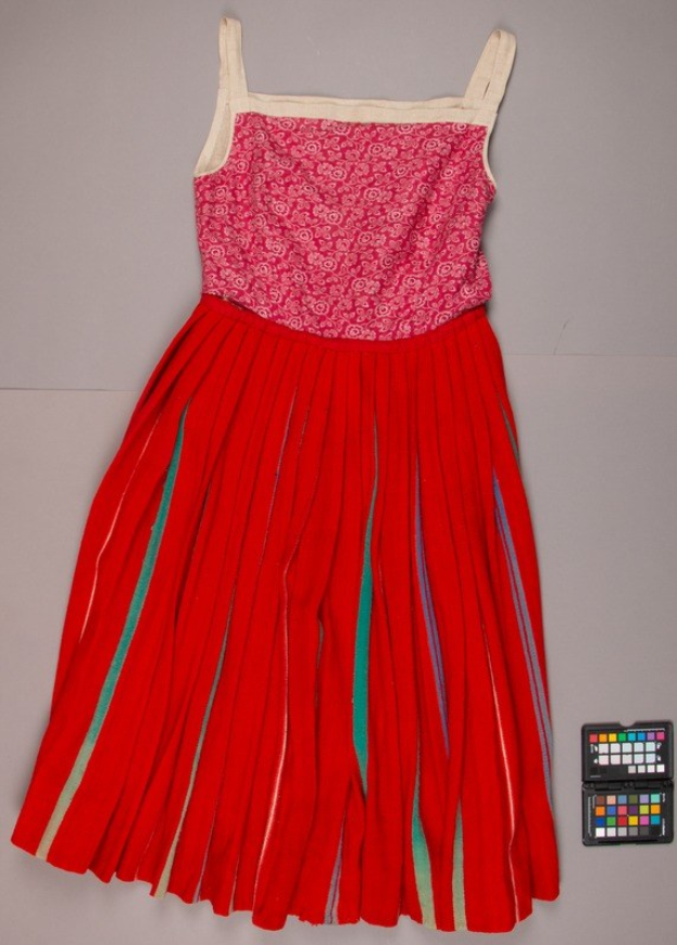 Ein rotes Kleid mit pinkem Oberteil und grünen Streifen, mit einem plissierten bodenlangen Rock, einem V-Ausschnitt und kurzen Ärmeln, das auf dem Boden neben einem leuchtenden Farbpaletten liegt.