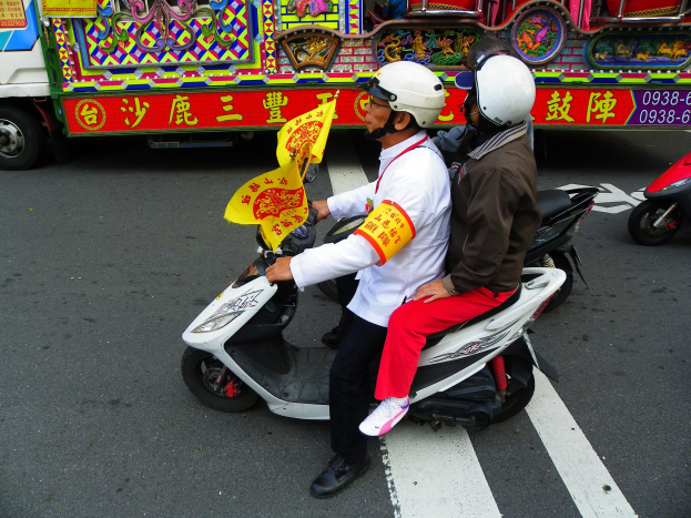 Zwei Personen mit Helmen fahren auf einem Motorroller, eine h"alt eine gelbe Flagge, mit einem Fahrzeug im Hintergrund.