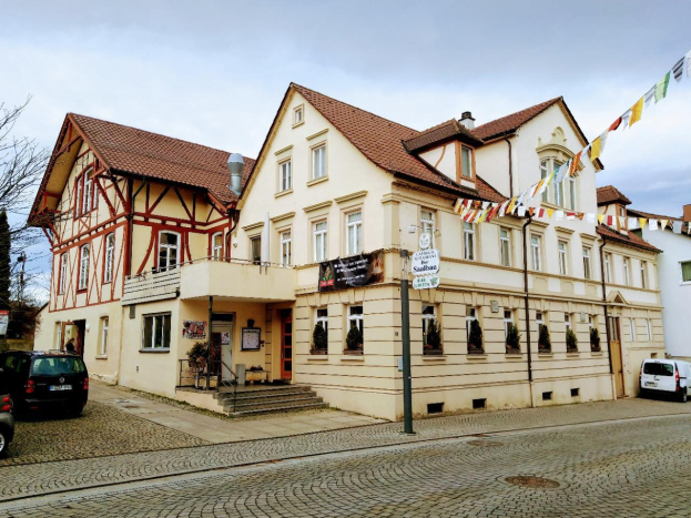 Das Stadtbahnhof-Gebäude in Baden-Württemberg, ein großer Bau mit zahlreichen Fenstern, ist von Bäumen, Straßenlaternen, Straßenlaternen, Kraftfahrzeugen, Fußgängern, Werbetafeln, Dekorationsfahnen und einem bewölkten Himmel umgeben.