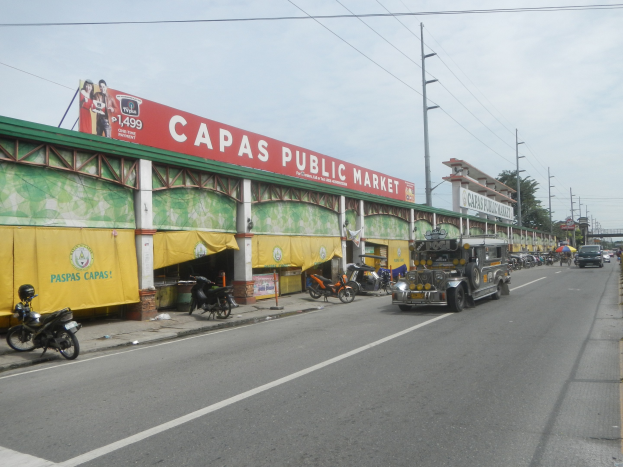 Eine belebte Stadtstraße mit Fahrzeugen, Fußgängern auf einem Gehweg, Strommasten, Gebäuden, Bäumen und einem bewölkten Himmel, mit einem Gebäude mit der Aufschrift "Capas Public Market" im Vordergrund.