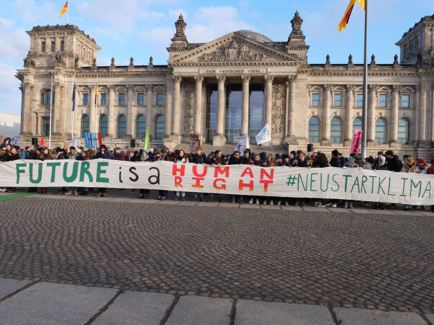 Gruppe von Menschen mit einem Banner 'Zukunft ist ein Mensch' vor dem Reichstaggebäude in Berlin, Deutschland, mit architektonischen Details des Gebäudes und Flaggen im Hintergrund bei bewölktem Himmel.