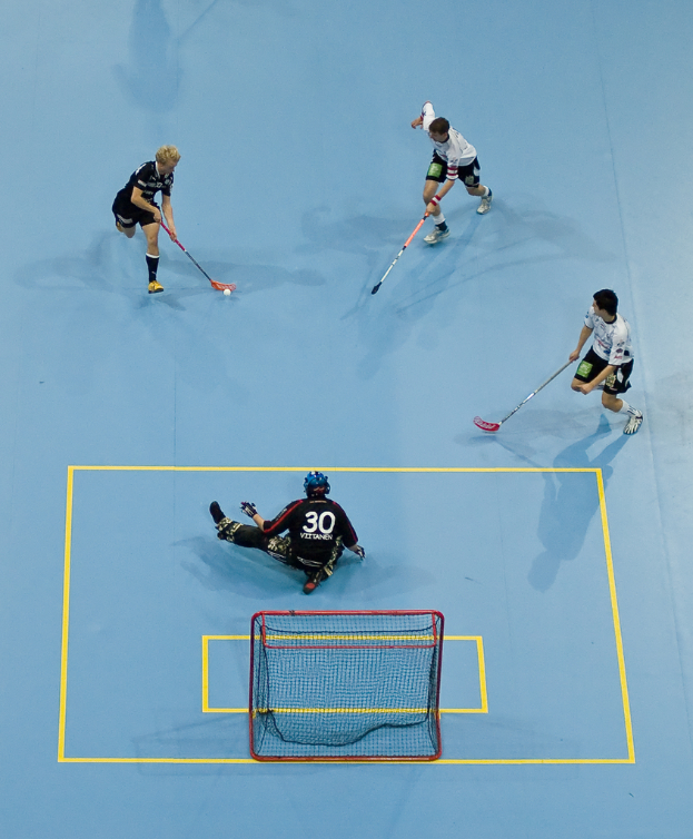 Drei Personen in Sportkleidung spielen Hockey, wobei eine Person rechts mit einem Hockeystock einen Ball schlägt und eine Person hinter einem Tor auf dem Boden sitzt.