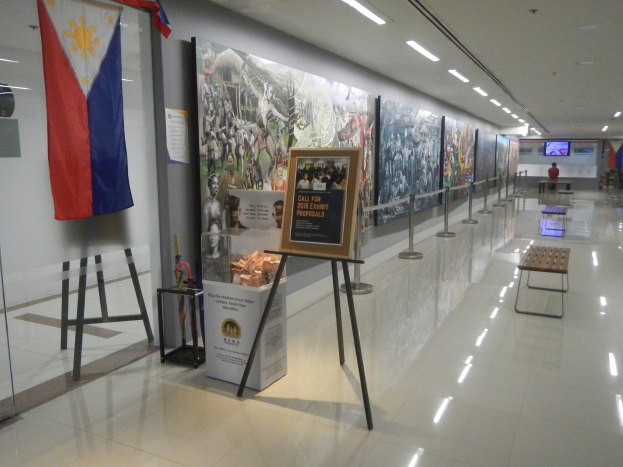 Langer Flur mit einer philippinischen Flagge an der Wand, Schautafeln mit Text und Bildern, Ribbon-Ständer, Bänke, Deckenleuchten und ein Bildschirm im Hintergrund mit Museums-Informationen.